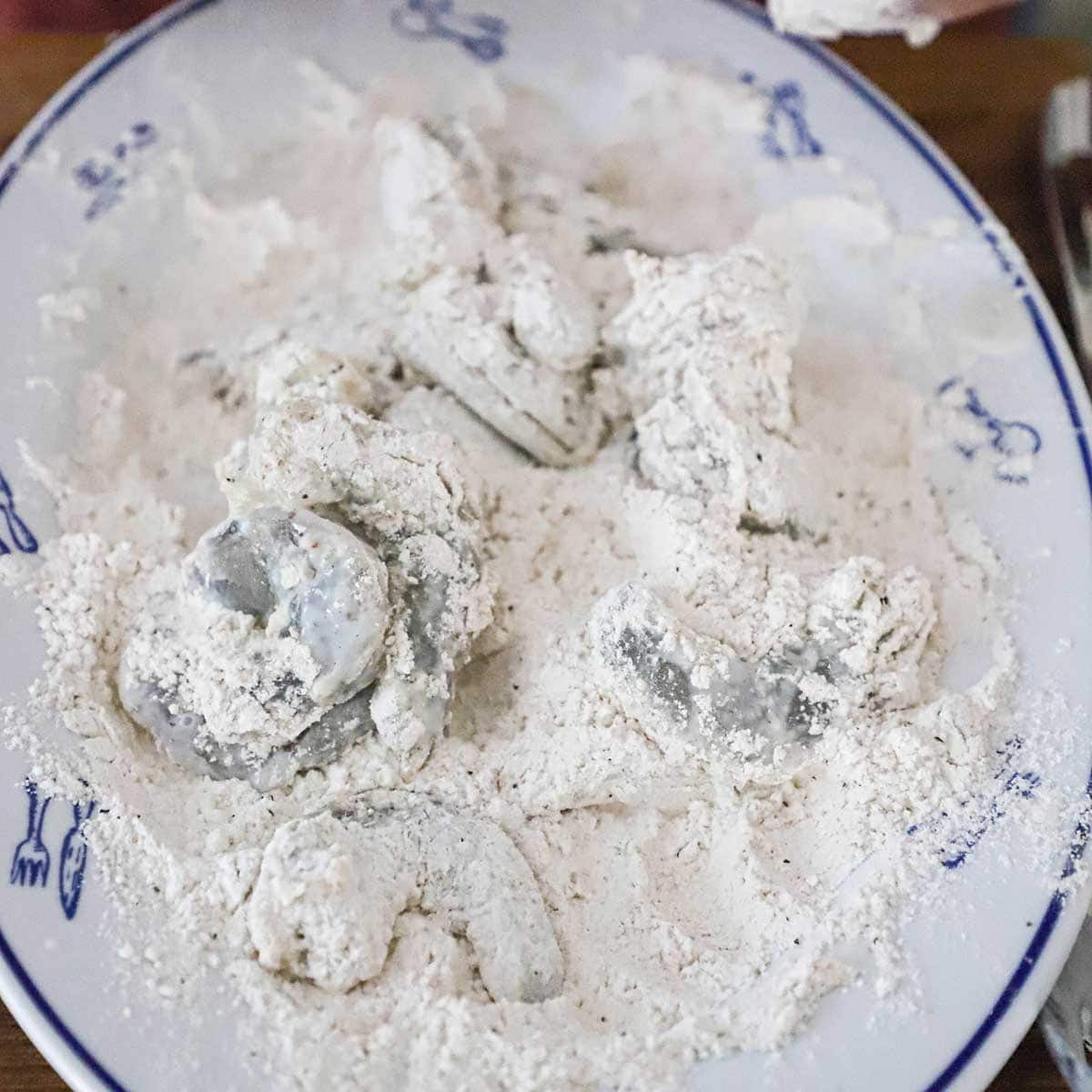 Uncooked shrimp that has been soaked in buttermilk being tossed in a platter filled with seasoned flour. 