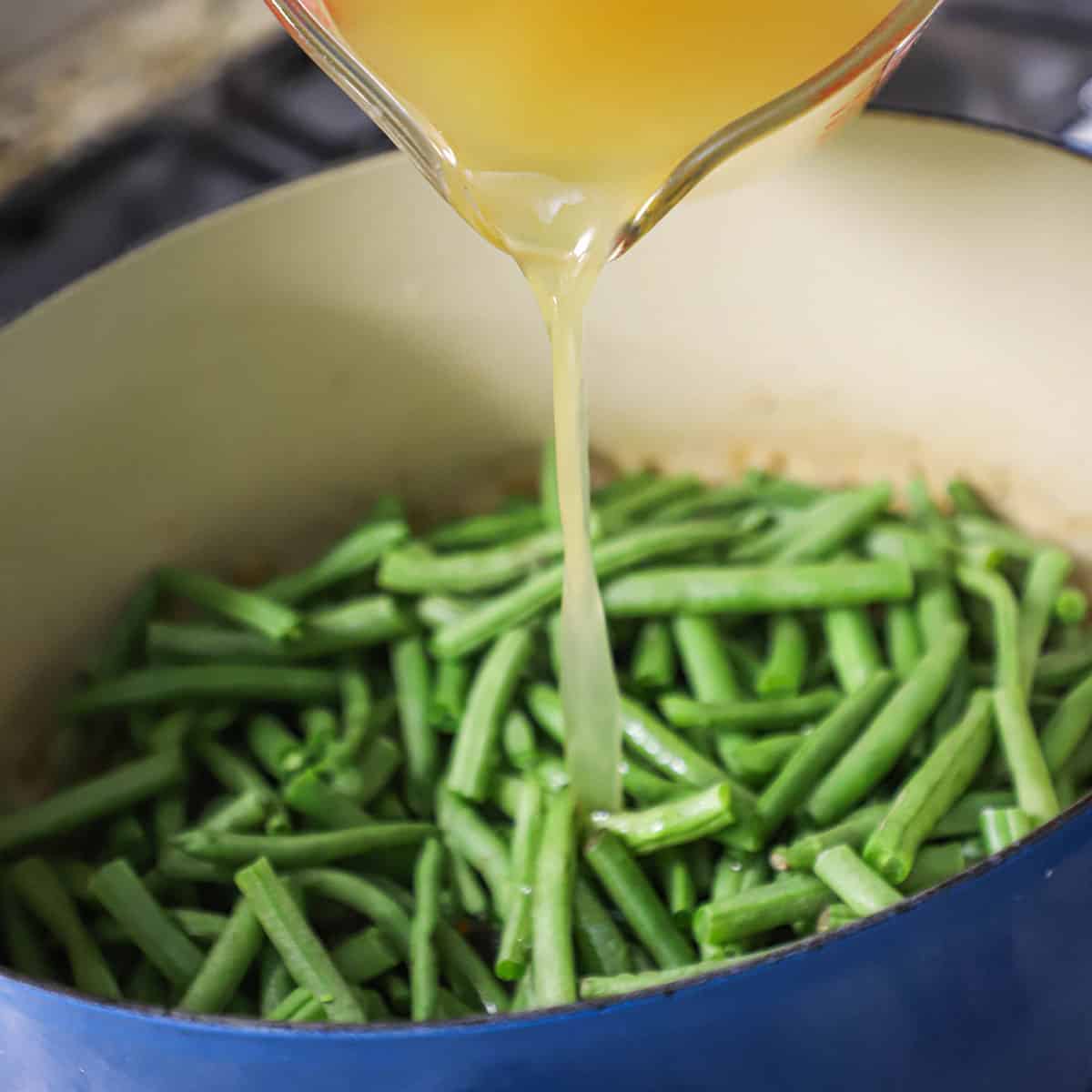 A person pouring chicken broth from a large glass measuring cup into a Dutch oven filled with fresh green beans.
