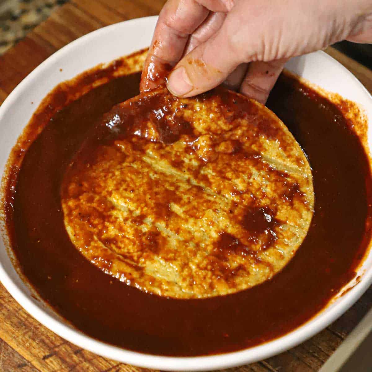 A person submerging a lightly fried corn tortilla into a bowl of red enchilada sauce. 
