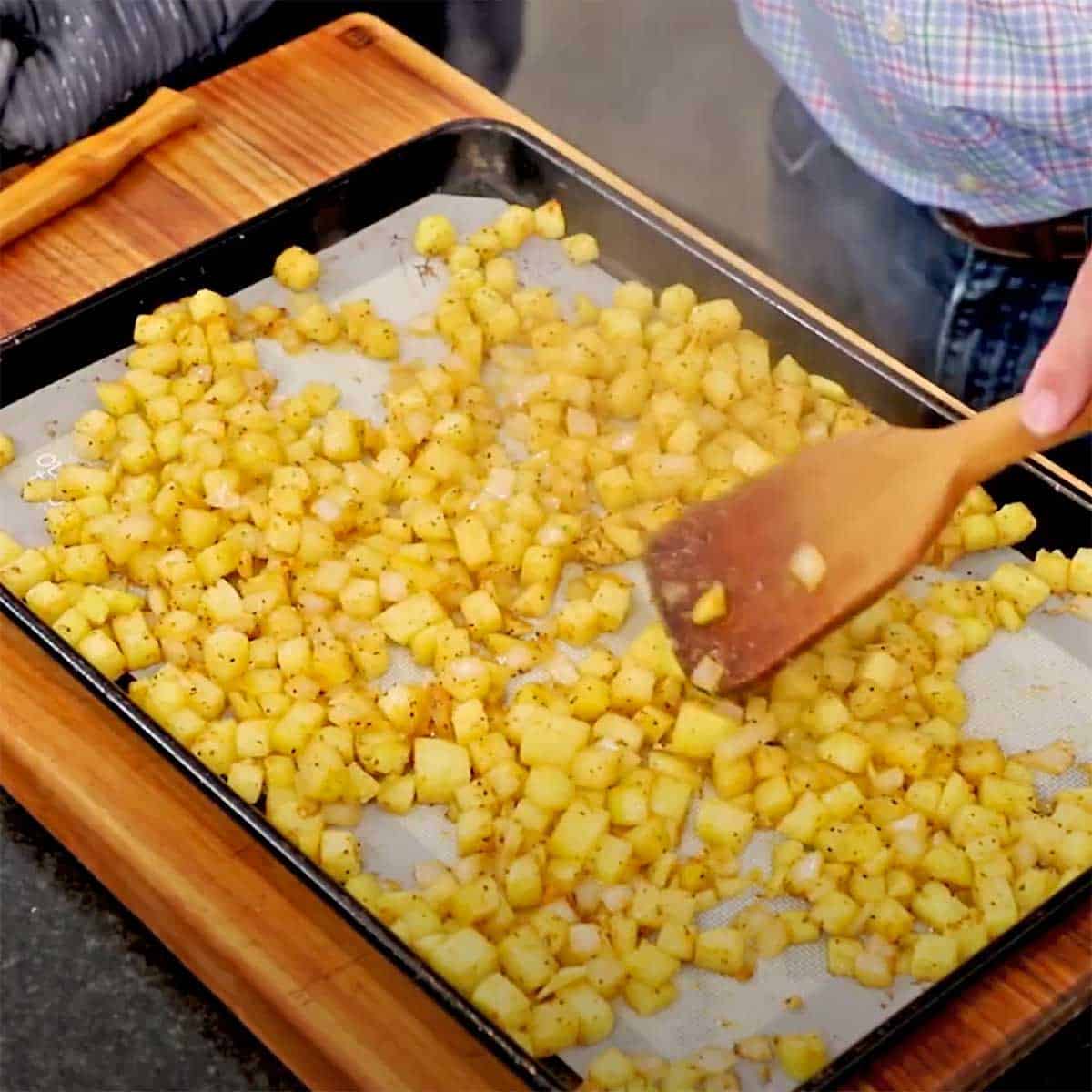 A person using a wooden spatula to move around partially cooked breakfast potatoes on a baking pan. 