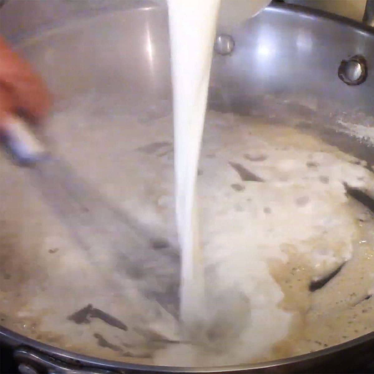 A person using a whisk to stir in milk into a roux in a large silver skillet. 