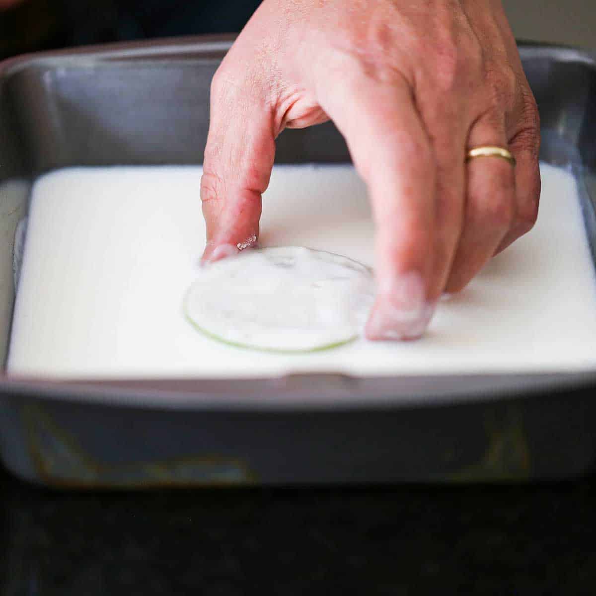 A person submerging a floured slice of green tomato into buttermilk that is in a square metal pan. 
