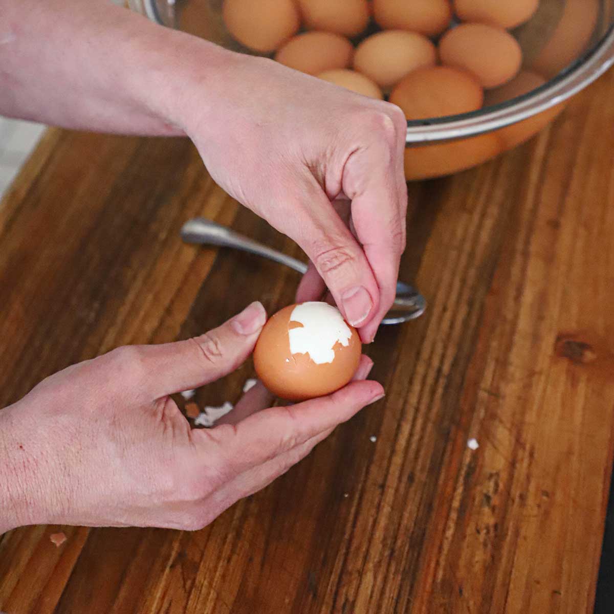 A person using his hands to peel the skin away from a hard boiled egg on a cutting board. 