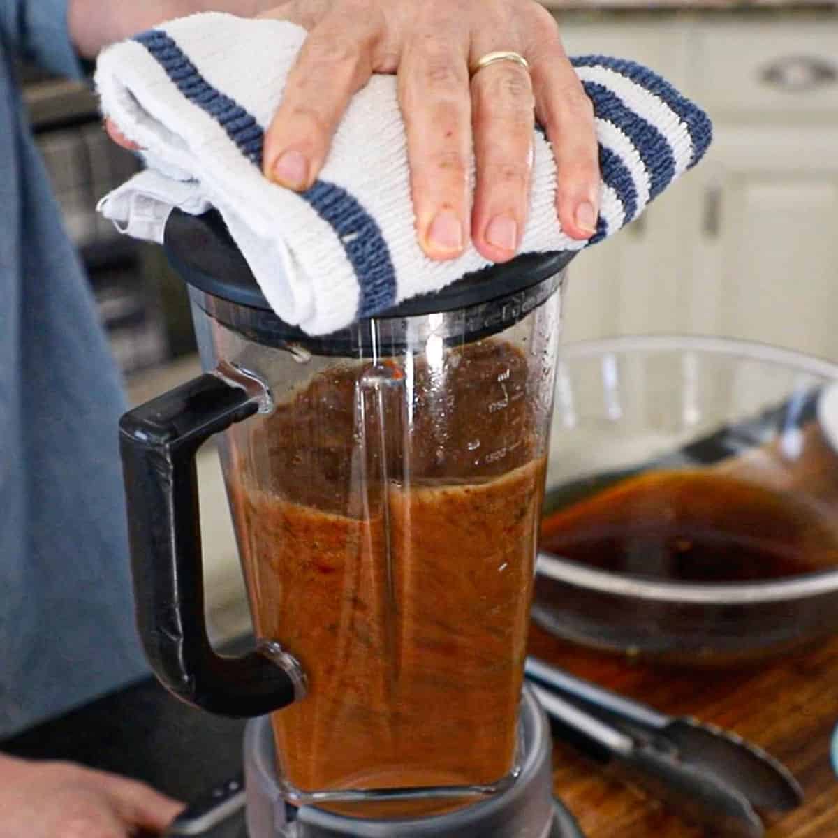 A person holding a kitchen towel over the top of a blender that is filled with puréed chiles, tomatoes, and onions. 