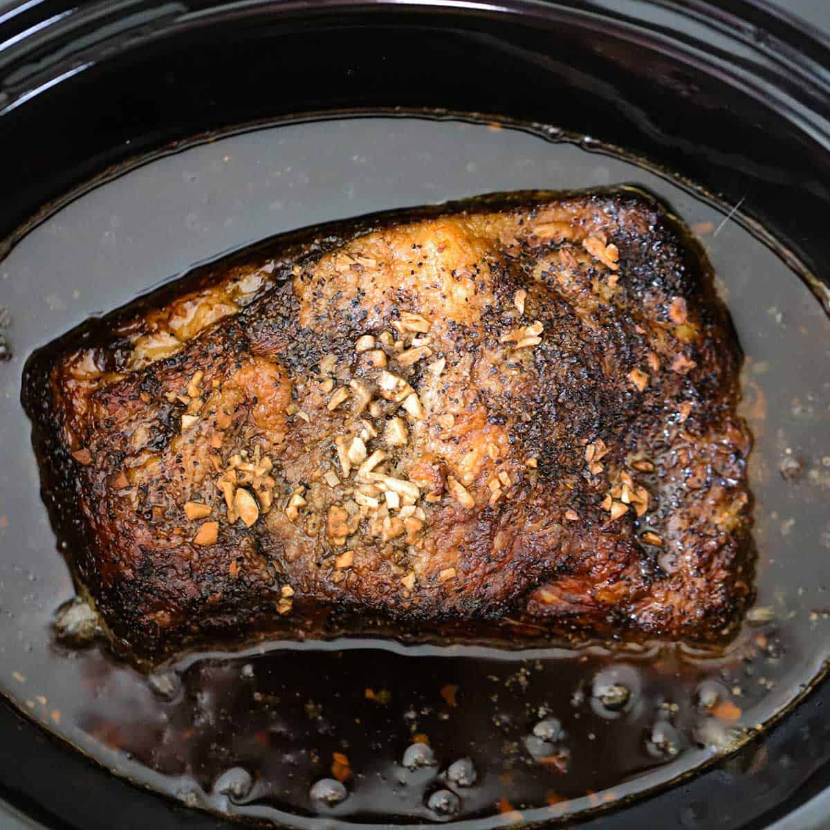 A fully cooked brisket resting in a thin, dark liquid in a black slow cooker.