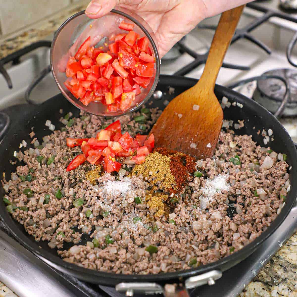 A person dumping chopped tomatoes from a small bowl into a large skillet filled with cooked ground beef, onions, and peppers.