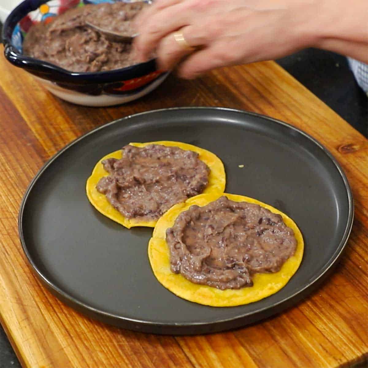 A person spreading homemade refried beans over two lightly fried corn tortillas that are resting on a large black dinner plate. 