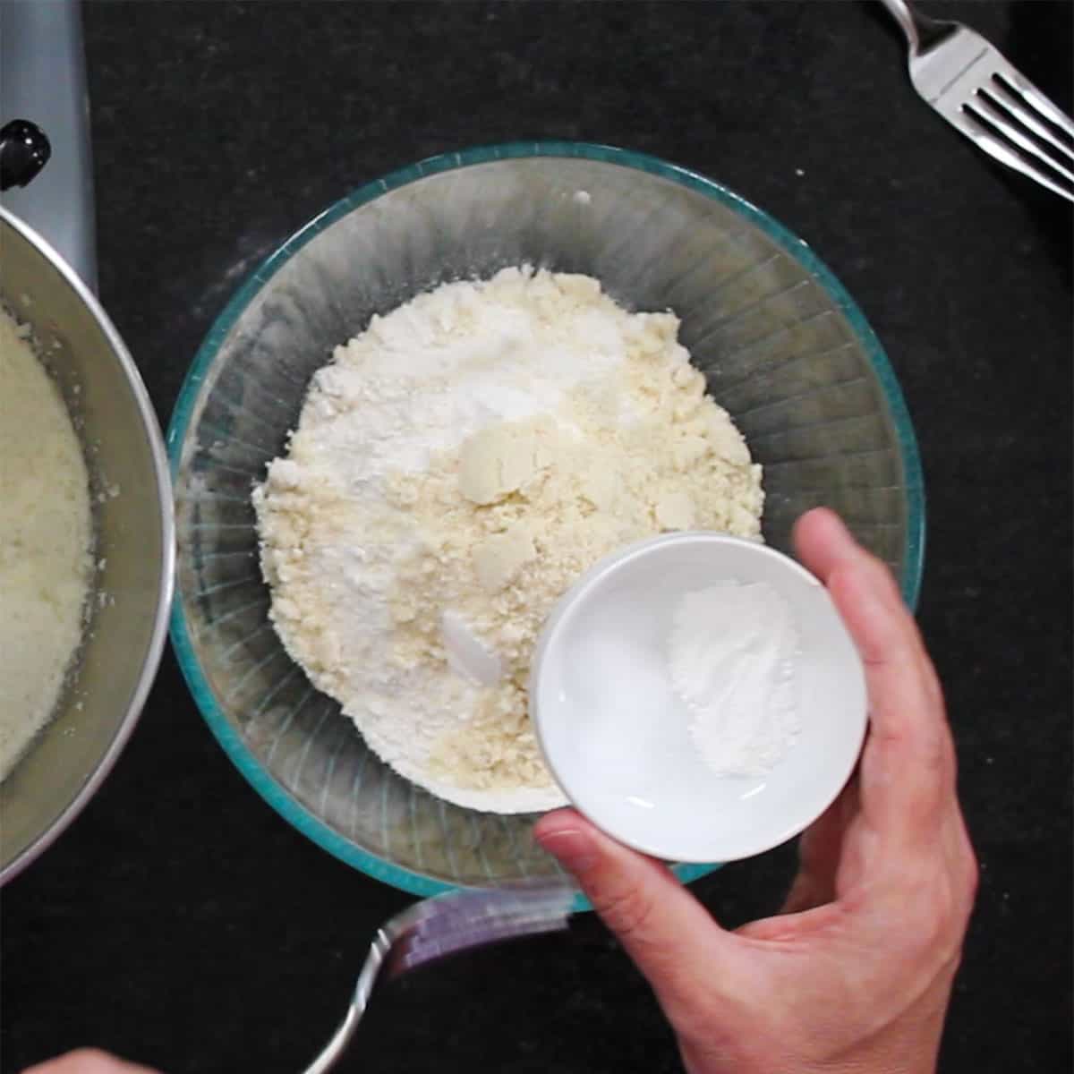 A person holding a small white bowl filled with baking powder over a glass bowl filled with all-purpose flour and almond flour. 