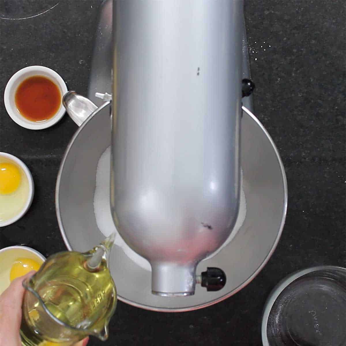 A person pouring vegetable oil into the bowl of a stand mixer that has sugar in it. 