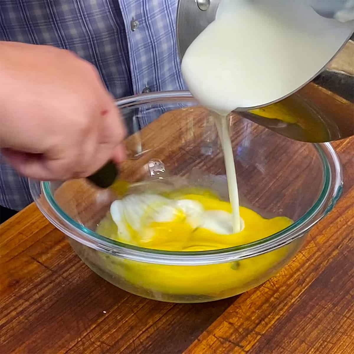 A person pouring a hot custard into a glass bowl filled with egg yolks. 