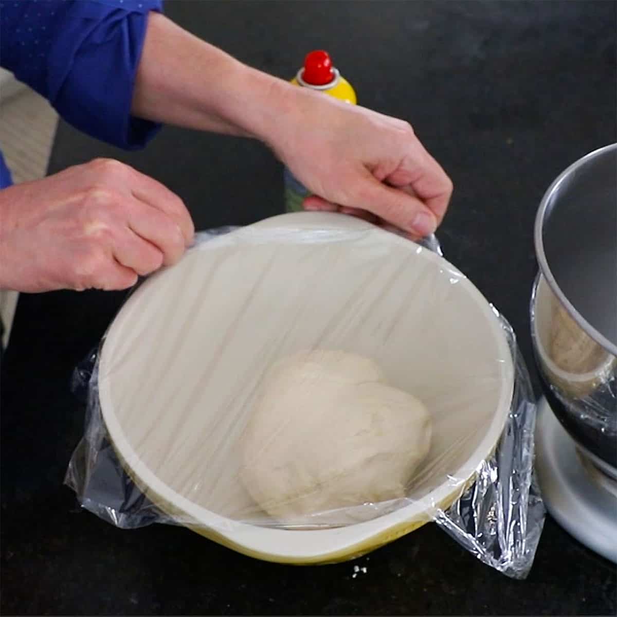 A person stretching plastic wrap over a ceramic bowl that is filled with a ball of un-risen dinner roll dough.