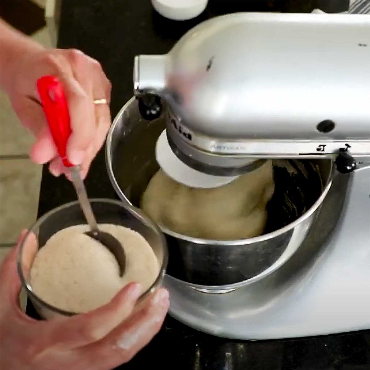 A person spooning in a foamy yeast mixture into a bowl of a mixer that has focaccia dough being mixed. 