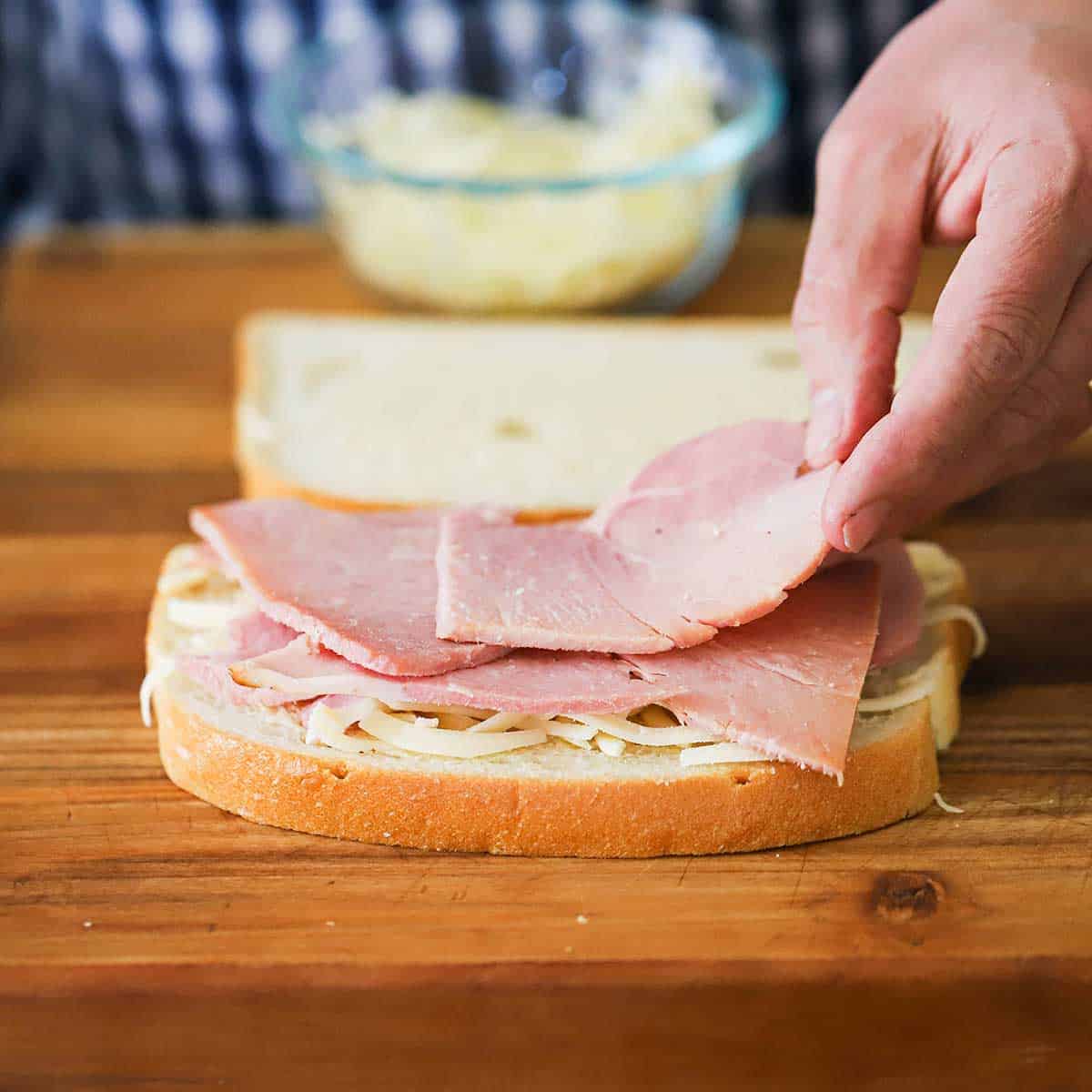 A person placing sliced ham on to a slice of bread that has a layer of grated cheese on it. 