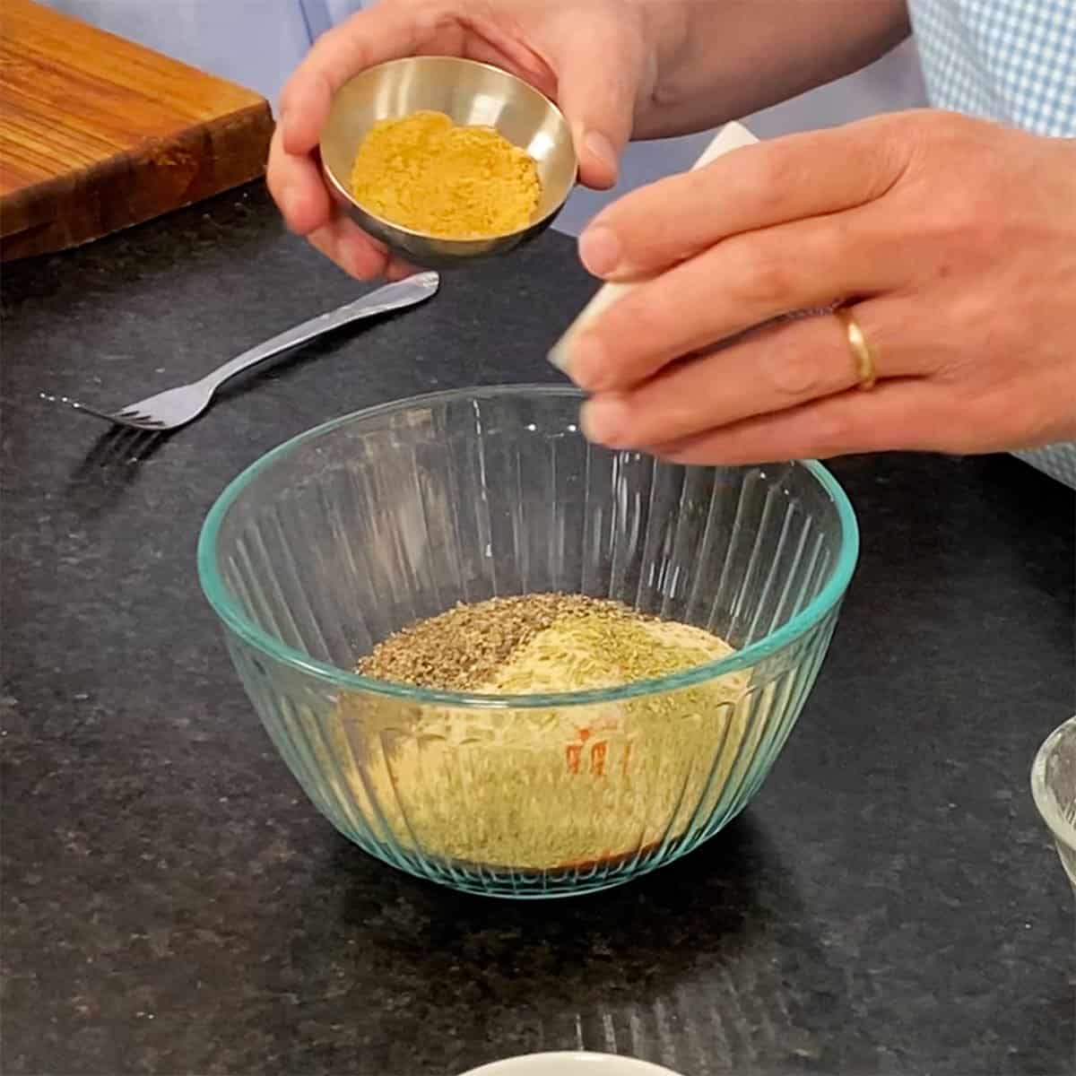 A person dumping ground ginger from a small metal bowl as well as dried rosemary from a small white bowl into a glass bowl filled with dried spices for pork rub.