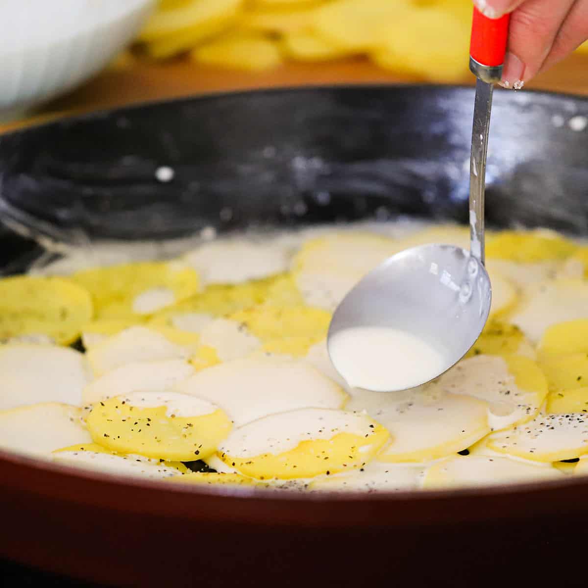 A person ladling a cream sauce over the tops of thinly sliced yellow potatoes in a large oval baking dish. 