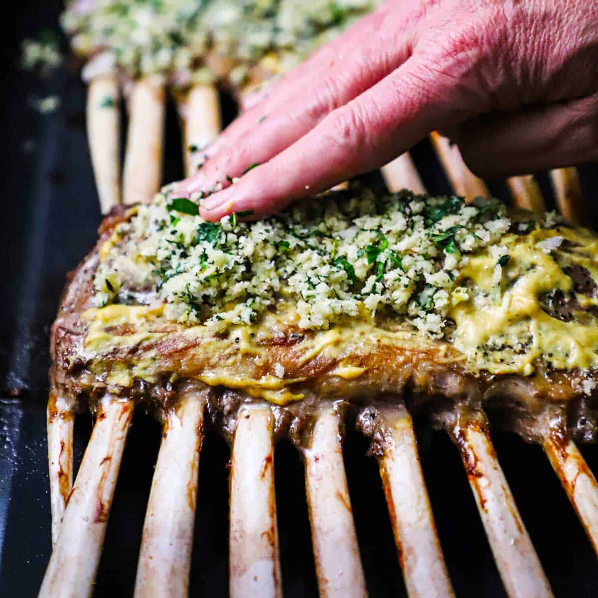 A person pressing a breadcrumb and herb mixture onto the top of a roasted rack of lamb.