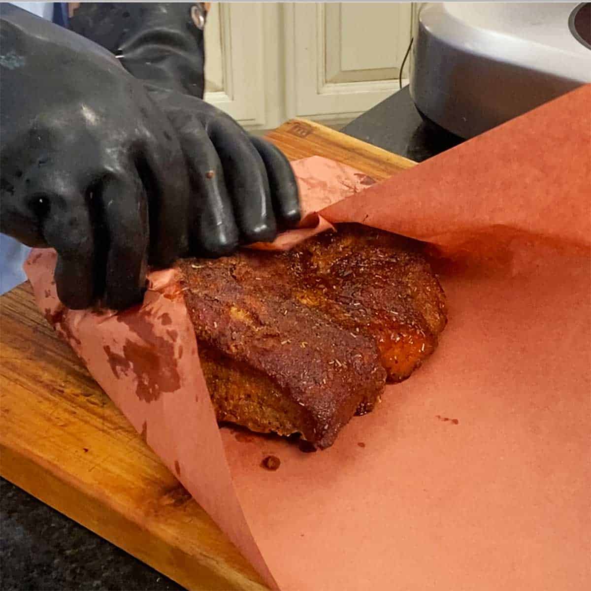 A person wearing heat-resistant gloves wrapping a partially smoked pork brisket in pink butcher paper. 