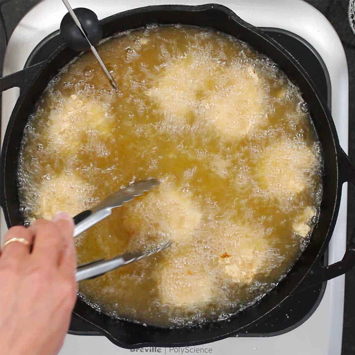 A person frying Southern shrimp in hot oil in a cast-iron skillet using a pair of metal tongs to turn the shrimp. 