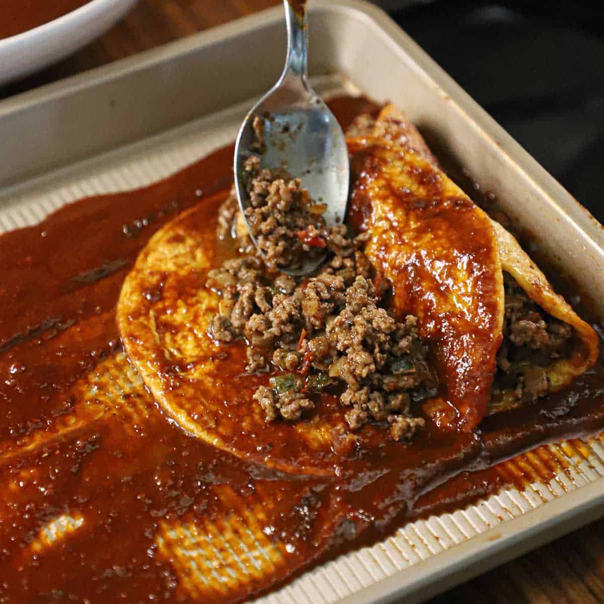 A person using a spoon to add a beef filling down the center of a corn tortilla that is coated in a red sauce. 