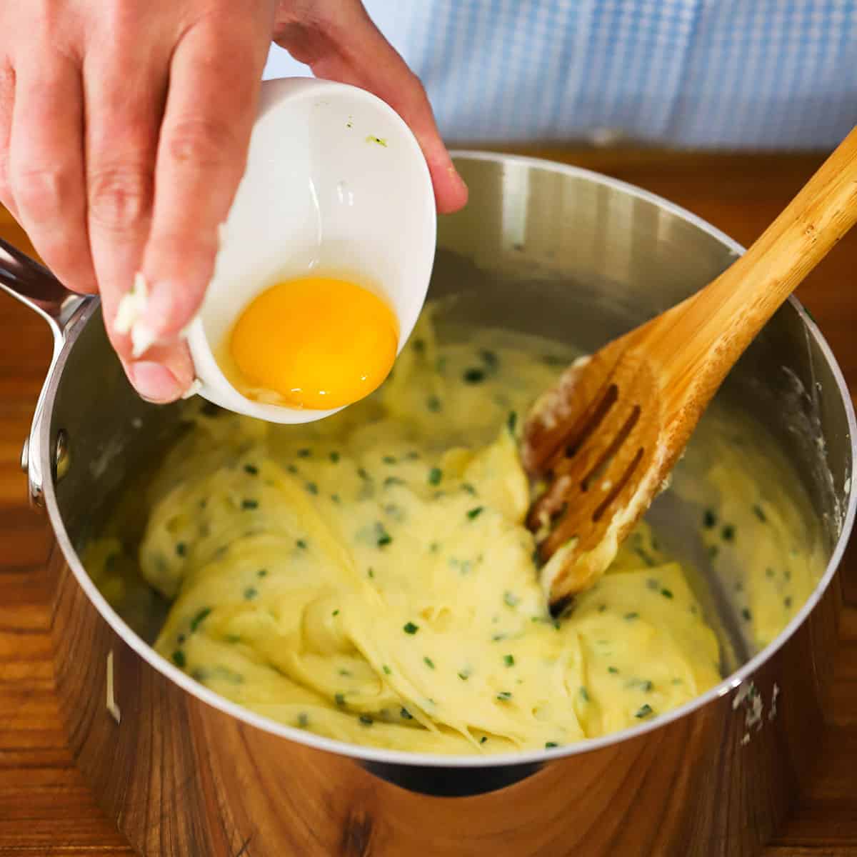 A person dumping an egg yolk from a small white bowl into a saucepan filled with the dough for a cheese soufflé with chives.