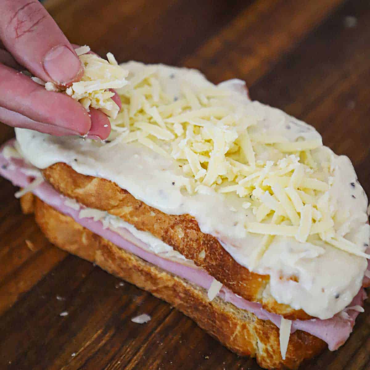 A person sprinkling grated Gruyere cheese over the top of a Croque Madama sandwich that is resting on a cutting board. 