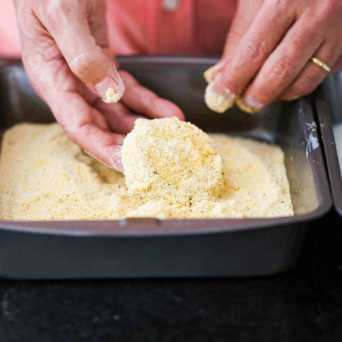 A person dredging a slice of green tomato through a cornmeal and breadcrumb mixture after it has been dredged in flour and then buttermilk. 