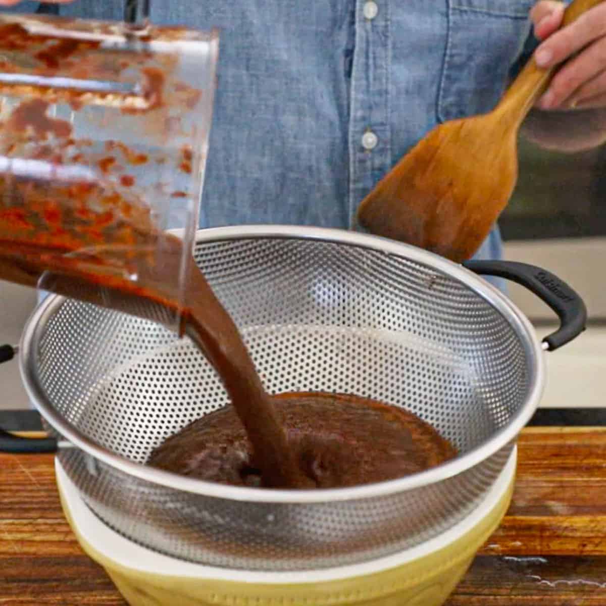A person pouring a puréed chili and vegetable liquid from a blender into a fine-mesh colander over a ceramic bowl. 