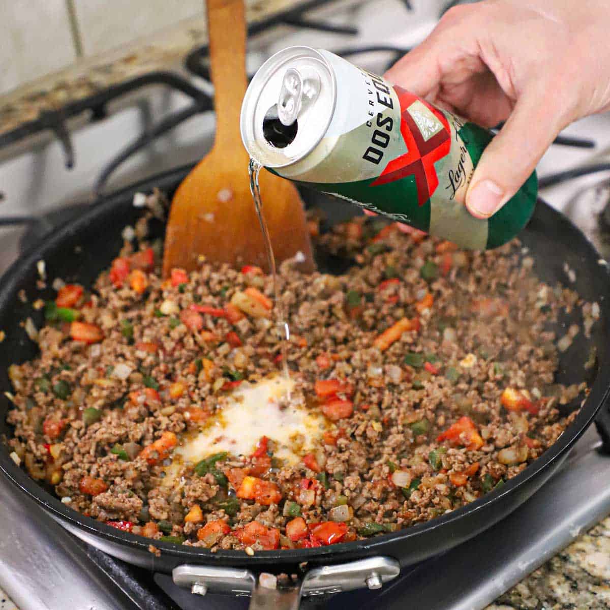 A person pouring beer into a skillet of simmering taco meat.