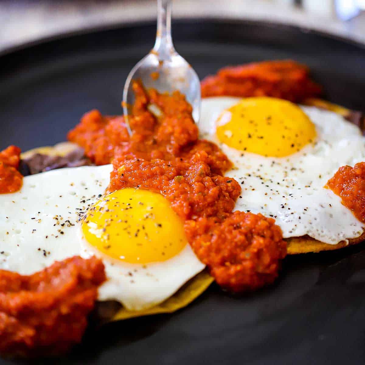 A person using a spoon to add homemade ranchero sauce over the tops of two fried eggs resting on refried beans and lightly fried corn tortillas all on a black dinner plate. 