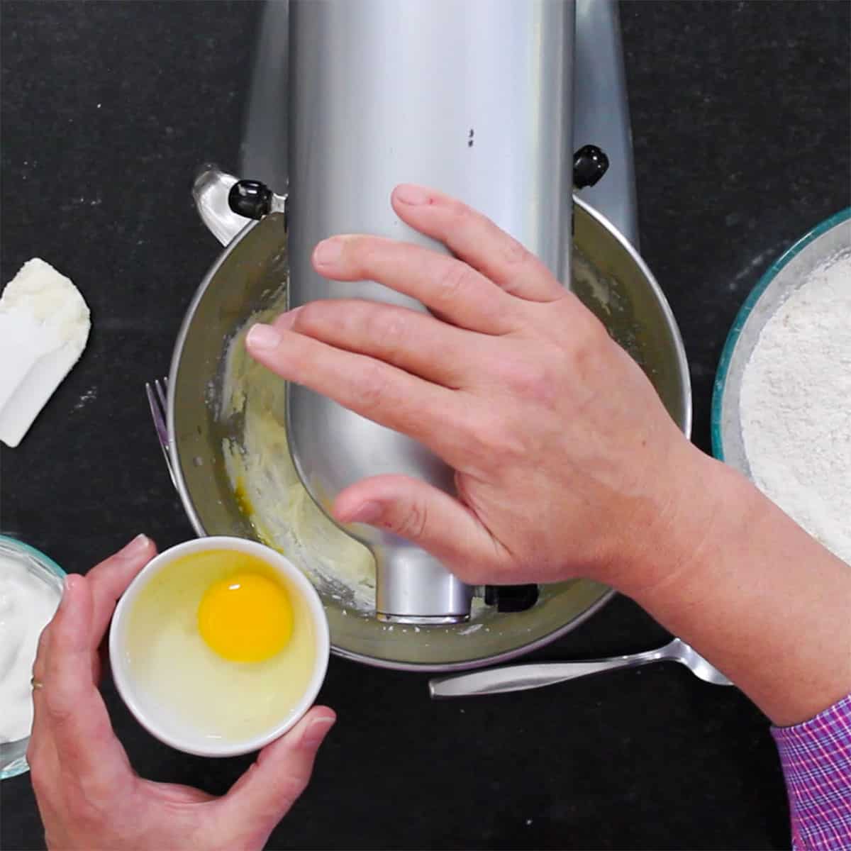 A person holding a small bowl that contains a whole egg over a stand mixer that is filled with a creamed butter and sugar mixture. 