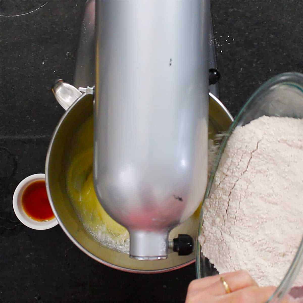 A person adding flour into the bowl of a stand mixer that already has eggs, oil, and sugar in it. 