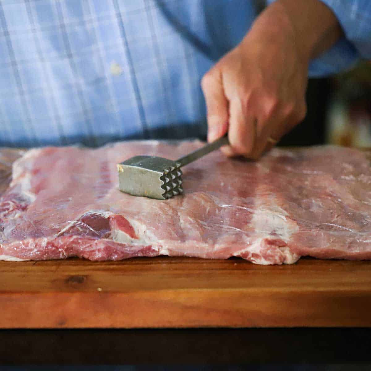 A person using the side of a meat mallet to pound a flat pork loin that is covered with plastic wrap. 