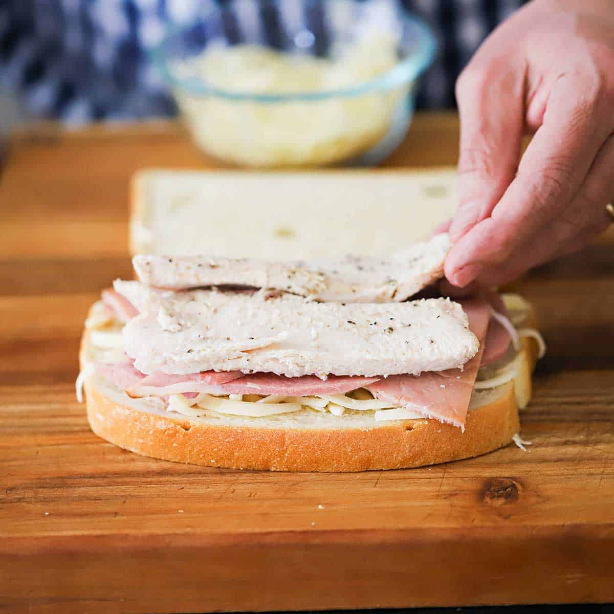 A person adding a layer of sliced roasted turkey on top of sliced ham on bread with grated cheese on it.  