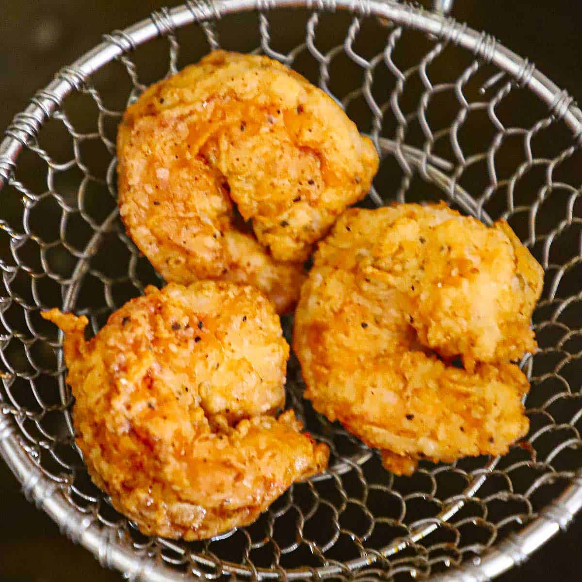 Three crispy Southern-fried shrimp sitting in a metal spider above a cast-iron skillet filled with hot oi. 