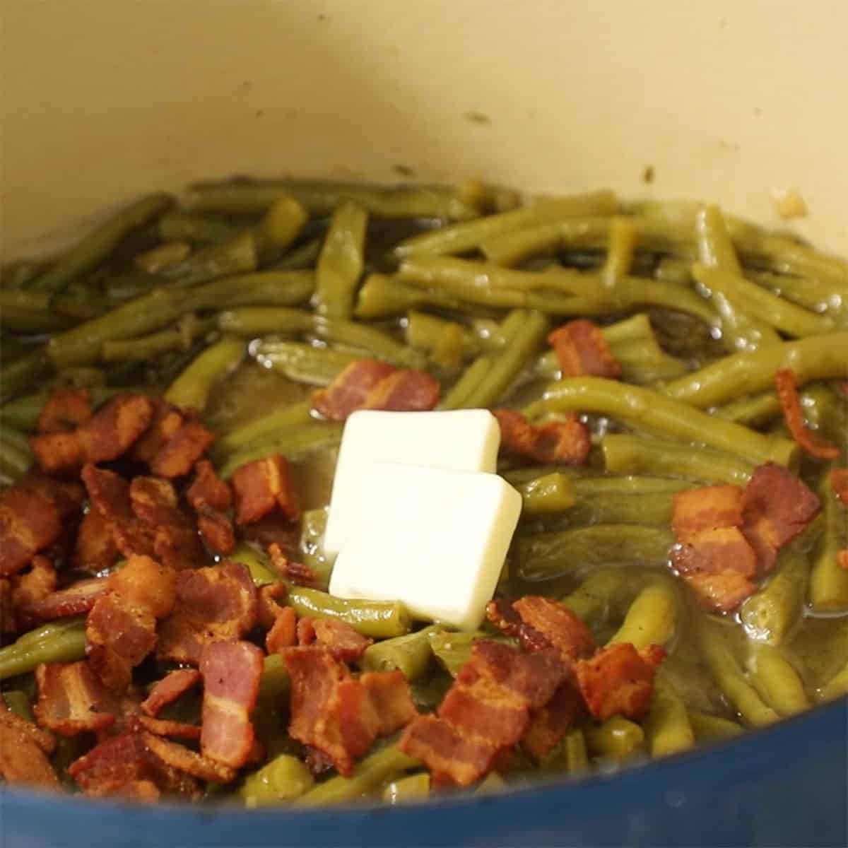 Two pads of butter and a pile of chopped crispy bacon on top of a pot of Southern-style green beans.