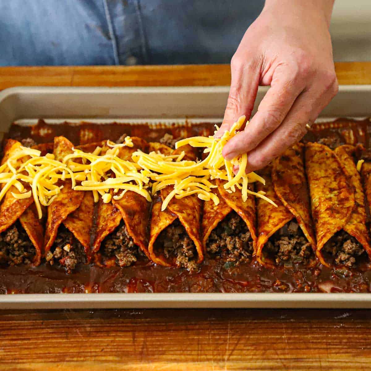 A person sprinkling shredded cheddar cheese over the top of beef enchiladas with red sauce. 