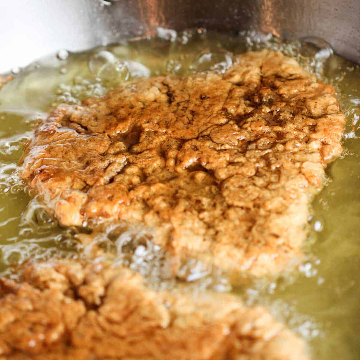 Two chicken fried steaks being fried in hot oil in a skillet. 