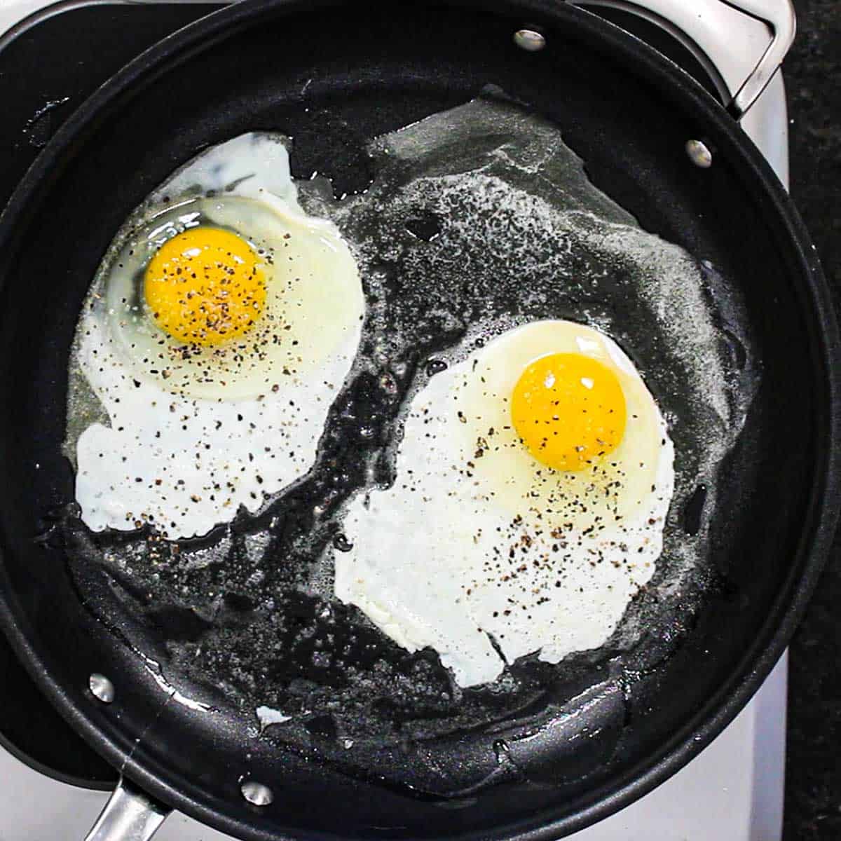Two eggs being fried in a large black skillet and have been seasoned with salt and pepper. 