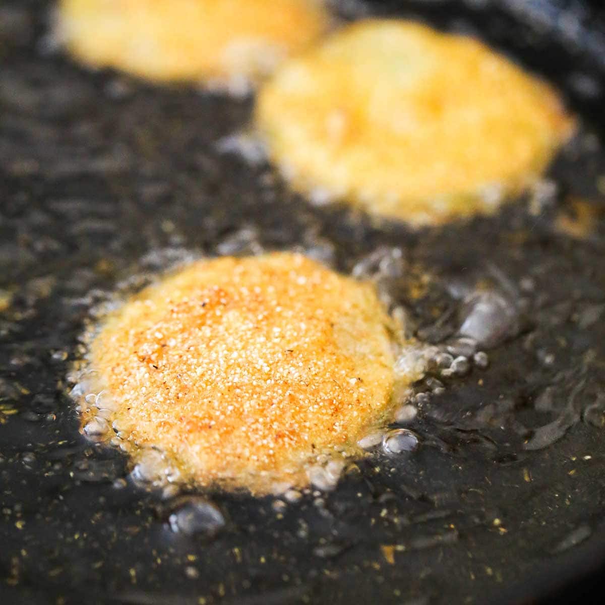 Several fried green tomatoes being deep fried in hot oil in a large cast-iron skillet. 