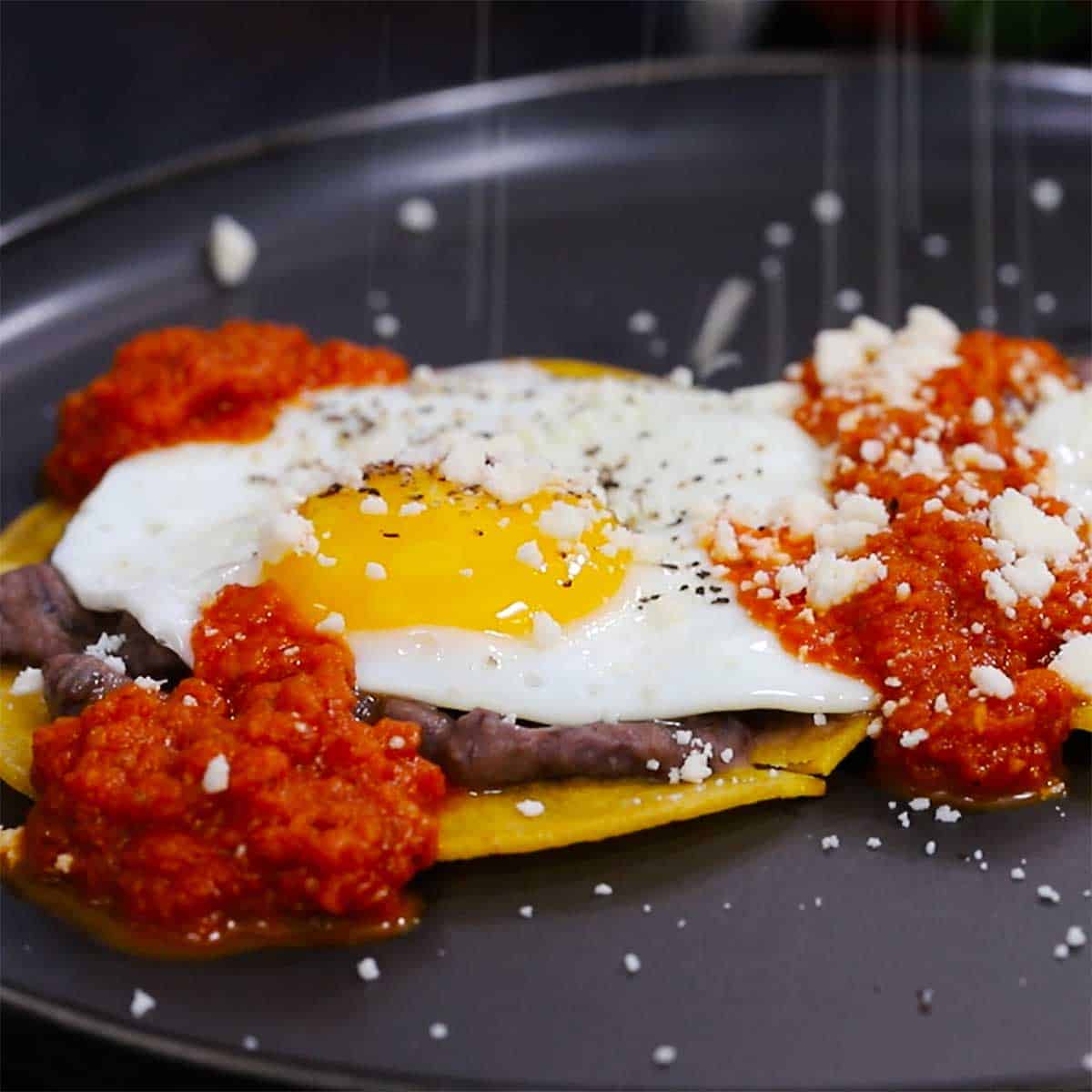 Crumbled Cotija cheese being sprinkled over the top of a plate filled with huevos rancheros. 