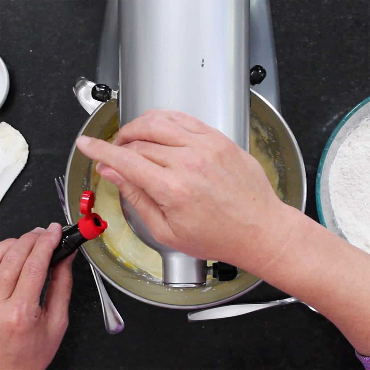A person pouring almond extract from a   small plastic container into the bowl of a stand mixer filled with creamed butter and sugar. 