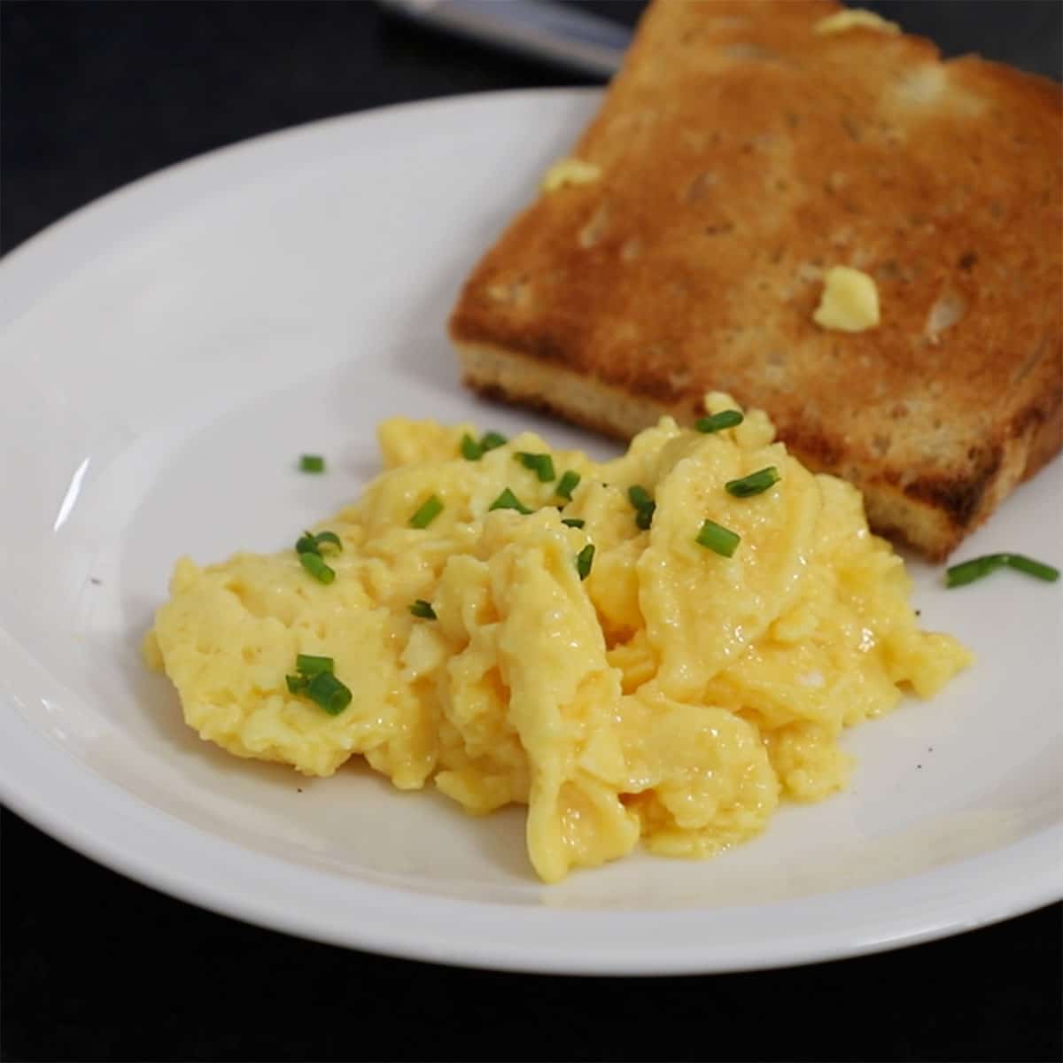 A white plate filled with a serving of fluffiest scramble eggs that are garnished with chopped chives with piece of buttered toast nearby. 