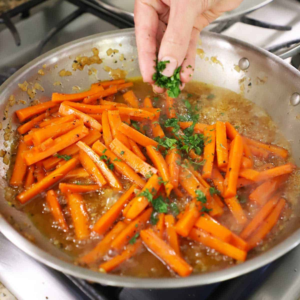 A person adding chopped fresh parsley into a skillet filled with simmering maple-glazed carrots.