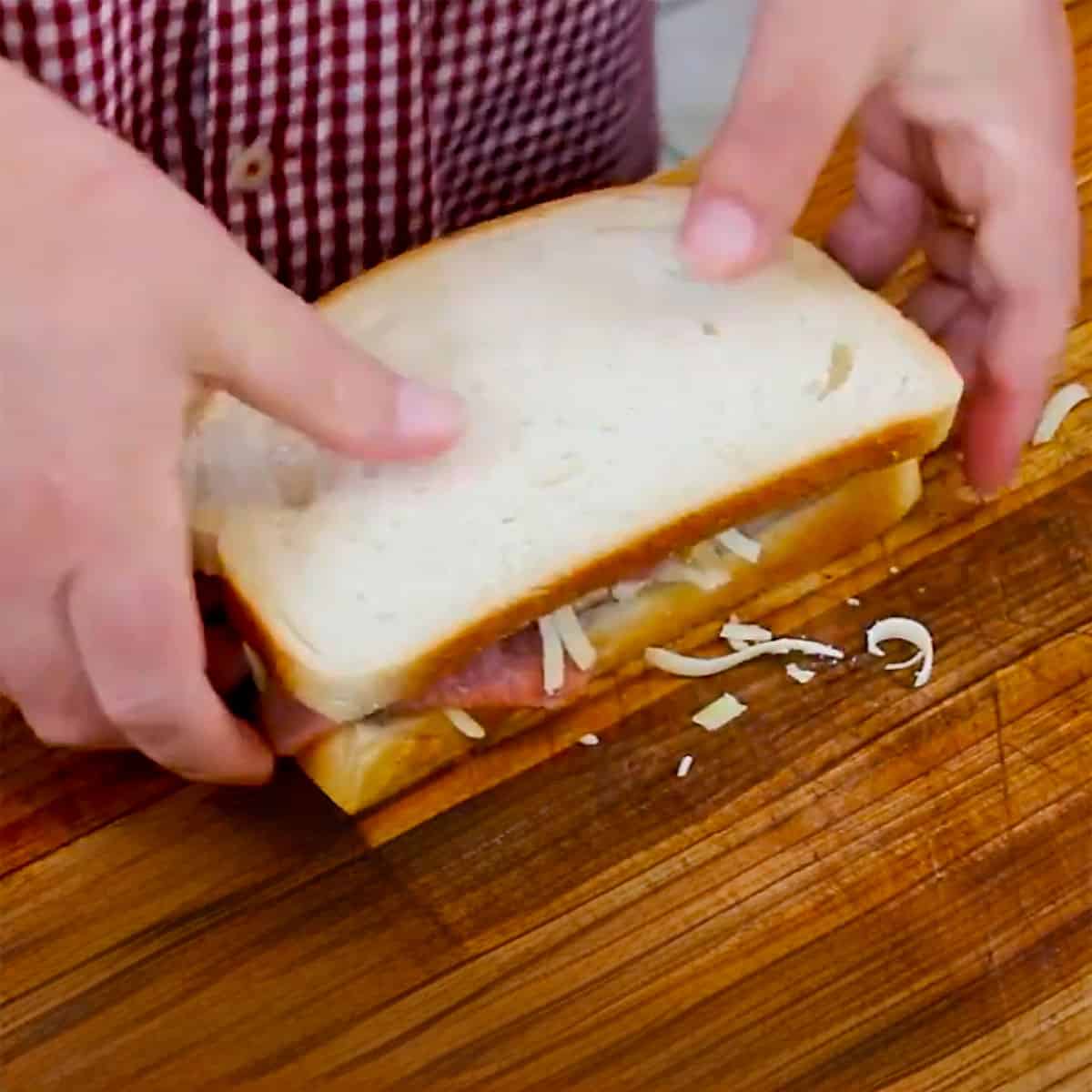 A person placing a top slice of sourdough bread on top of slices of ham and turkey and grated cheese on another slice of bread. 