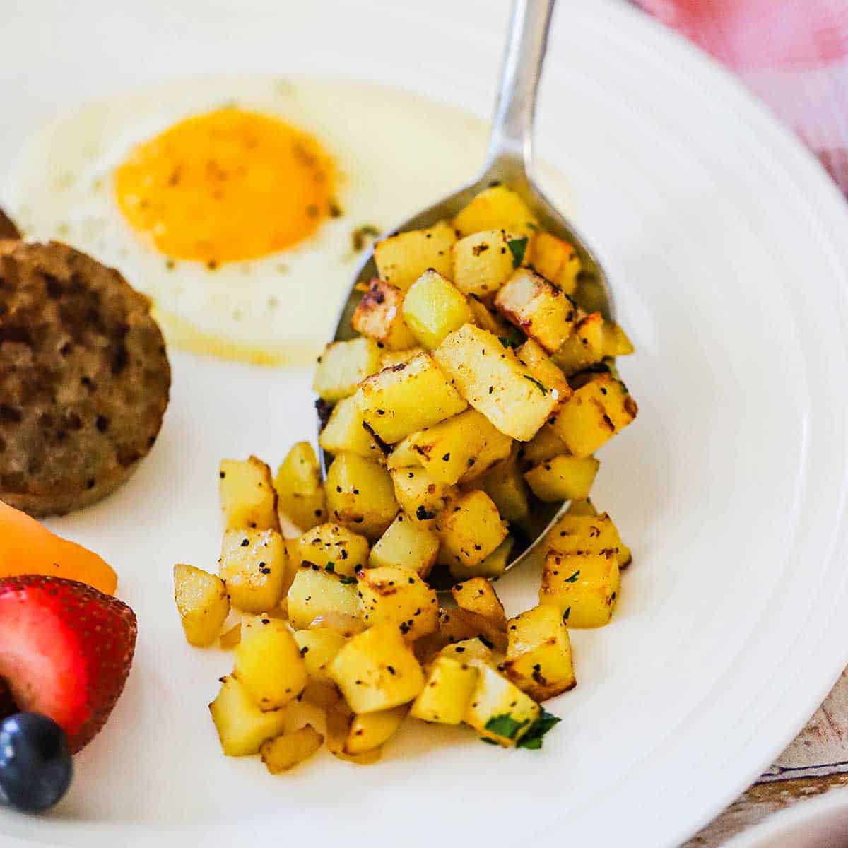 A person using a large serving spoon to add cooked breakfast potatoes onto a white plate next to a fried egg and fresh fruit. 
