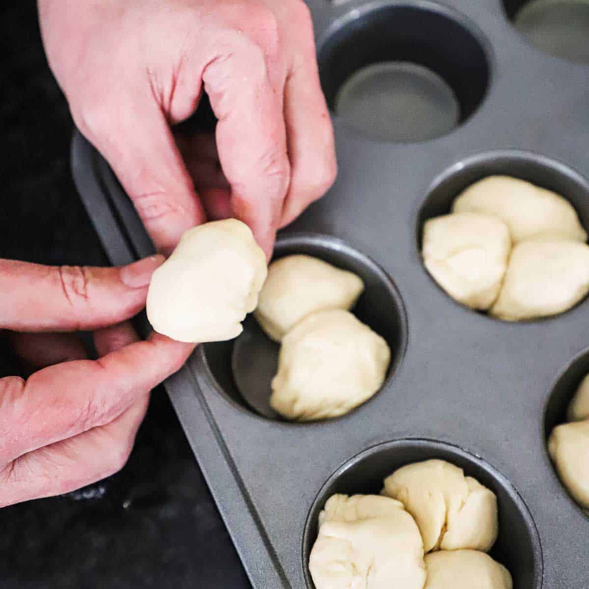 A person placing three small ball of dough into the openings of a muffin pan. 