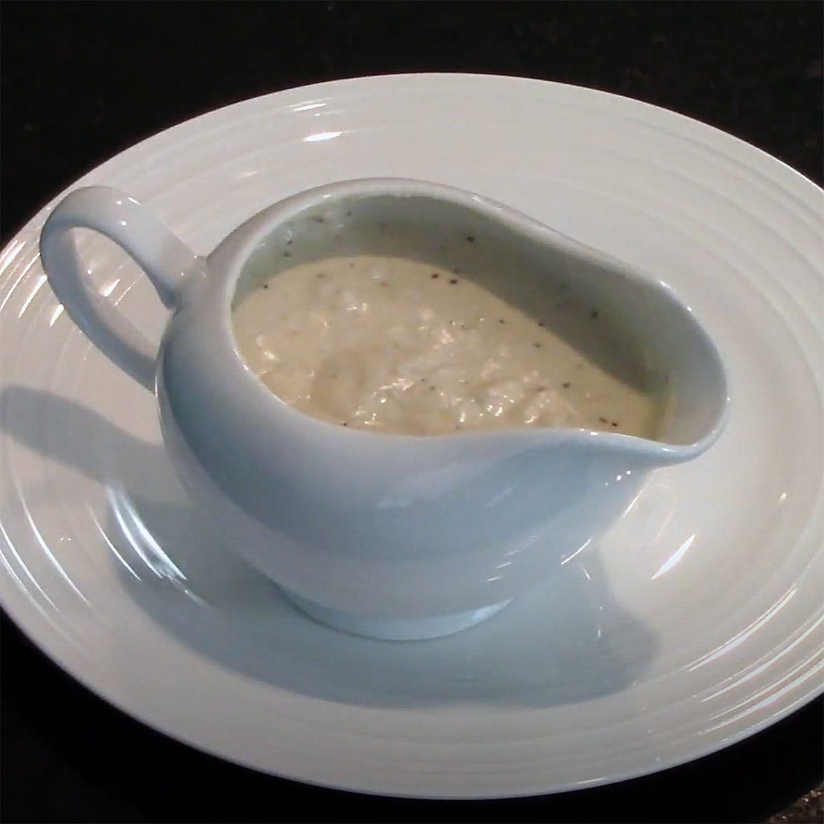 A white ceramic gravy boat filled with cream gravy on a white dinner plate. 