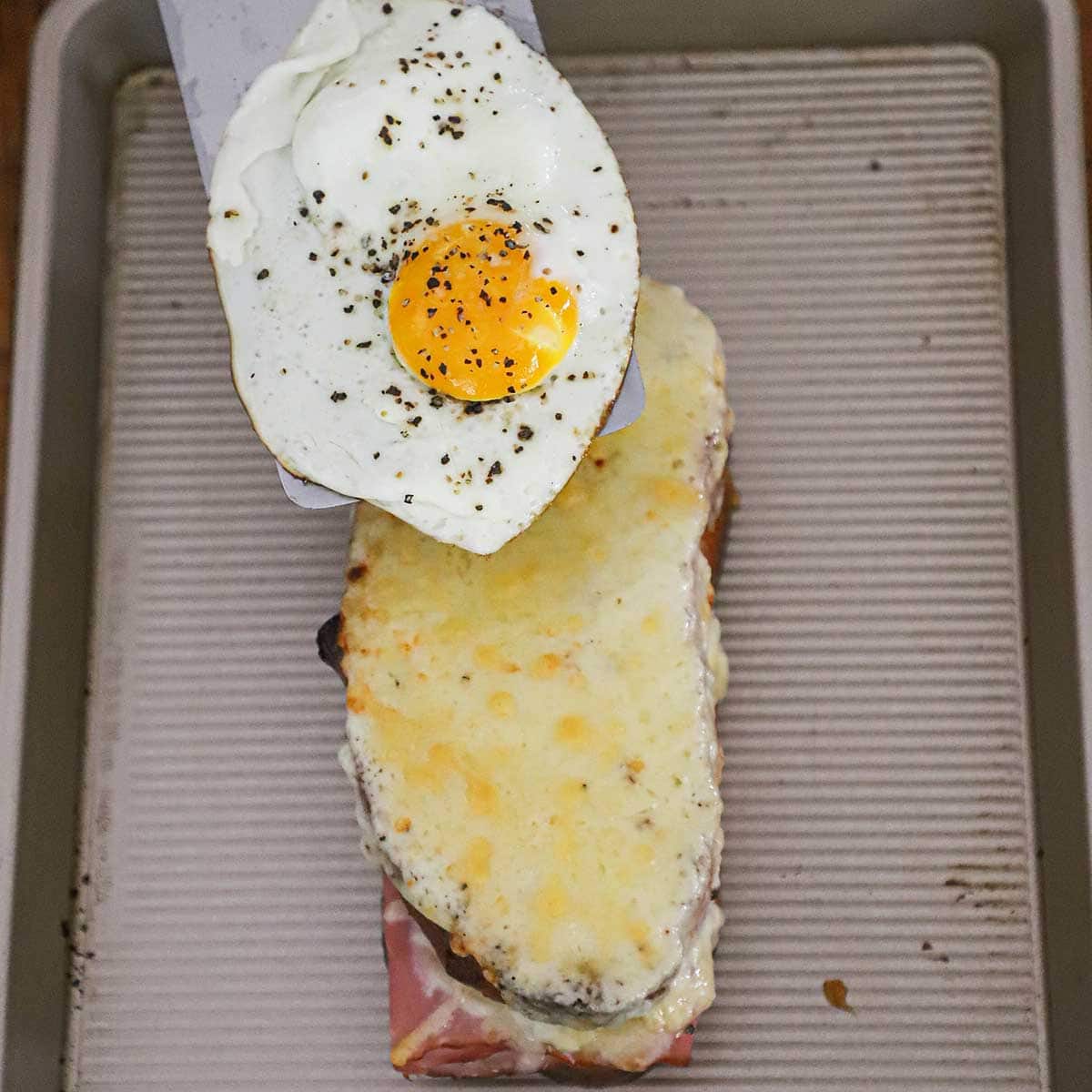 A person transferring a sunny side up fried egg from a metal spatula to the top of a baked Croque Madame sandwich. 