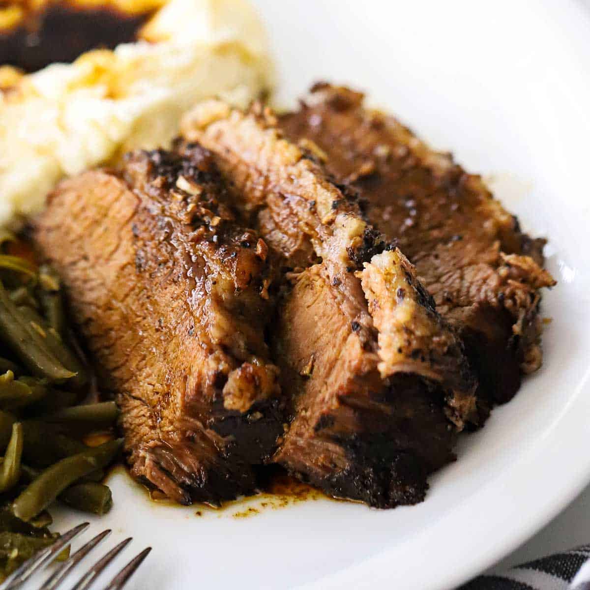 Three slices of slow-cooker brisket resting on a white dinner plate next to helping of green beans and mashed potatoes all topped with an au jus sauce.
