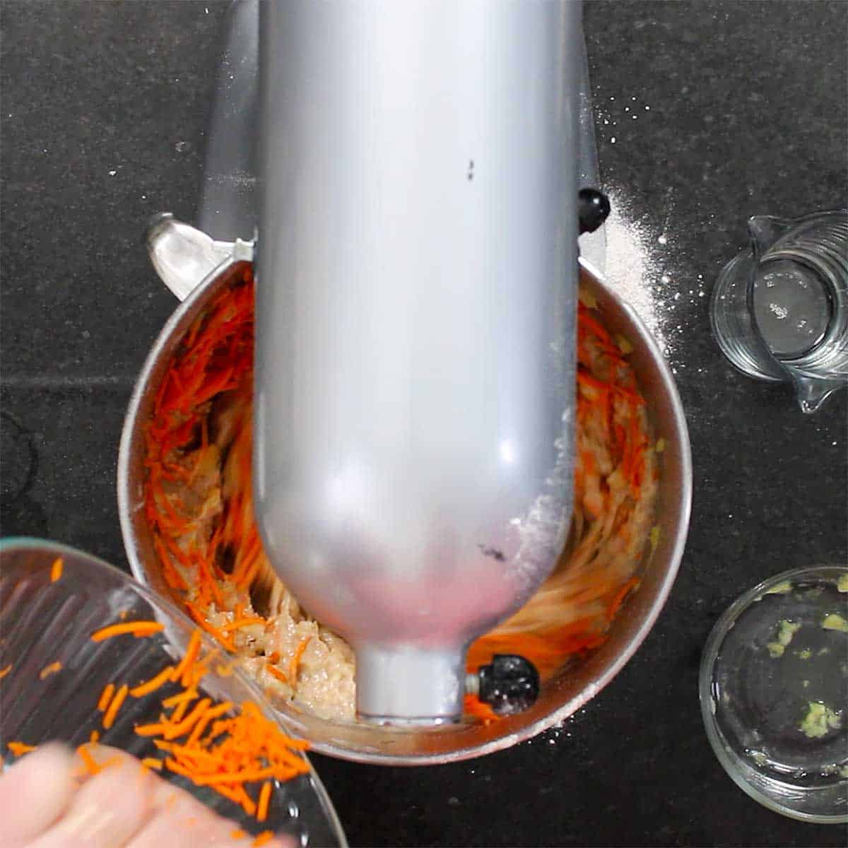 A person adding shredded carrots into the bowl of a stand mixer that is filled with a carrot cake batter. 